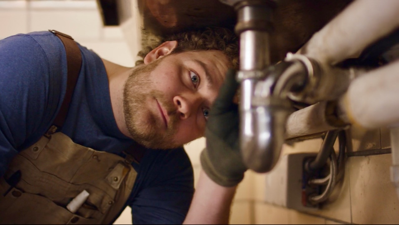 Plumber lying under a sink, looking up at exposed pipes, wearing blue shirt and beige overalls with a tool in the pocket.